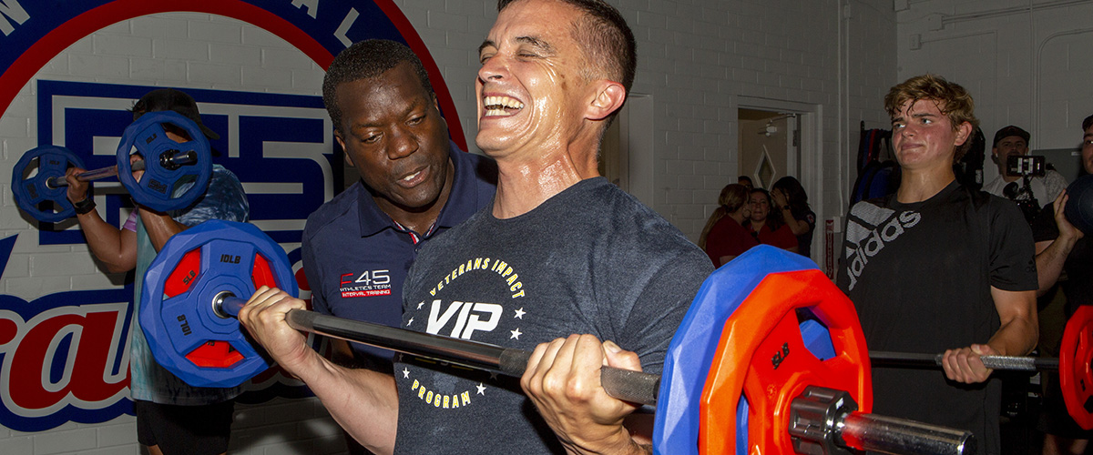 Image of mad curling a barbell, with a fitness trainer motivating him by his side.